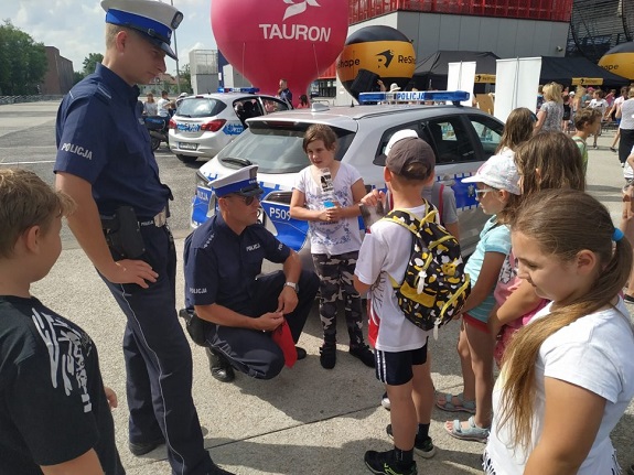 Dwaj policjanci w trakcie rozmowy z dziećmi podczas festynu. W tle widoczne radiowozy i motocykl.