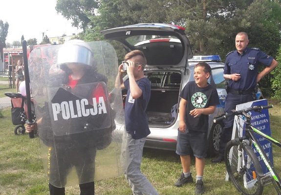 Na tle radiowozu stoi troje dzieci. Jedno z nich ma założony na głowę kask, a w ręku trzyma tarczę policyjną. Obok dzieci stoi policjant.