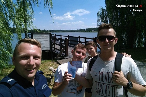 Na zdjęciu na pierwszym planie widać trzy osoby i policjanta na tle jeziora.
