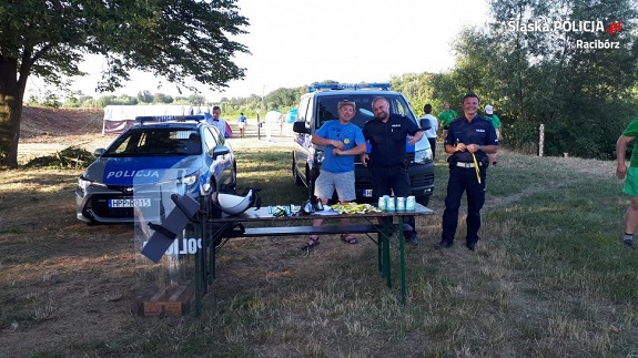 Na zdjęciu na drugim planie widoczne jest stoisko policyjne, na którym ustawione są dwa radiowozy. Natomiast na pierwszym planie widać policjantów w trakcie rozmowy z mężczyzną.