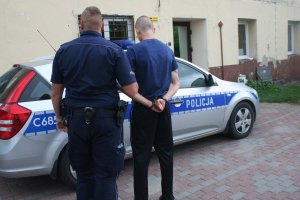 Policjant prowadzi zatrzymanego mężczyznę. W tle znajduje się policyjny radiowóz.