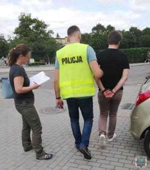 Zdjęcie przedstawia trzy osoby. Policjanta prowadzącego zatrzymanego mężczyznę. Obok stoi kobieta.