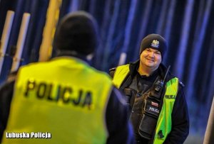 Na zdjęciu znajdują się dwaj policjanci. Jeden stoi z kamizelką odblaskową, drugi jest odwrócony.