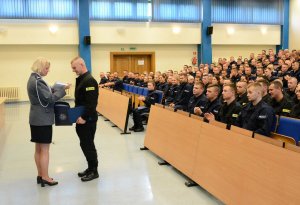 Uroczyste zakończenie szkolenia zawodowego podstawowego. Policjant odbiera dyplom ukończenia kursu. W tle widoczni są policjanci na auli.