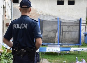 Na pierwszym planie znajduje się policjant, który stoi tyłem. W tle widać budynek oraz trampolinę.