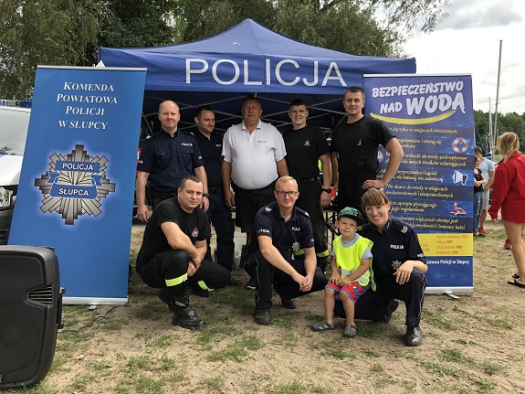 Na zdjęciu widoczna jest grupa osób - funkcjonariuszy Straży Pożarnej oraz Policji na tle Policyjnego namiotu.