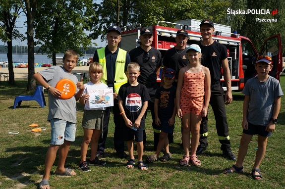 Zdjęcie przedstawia grupę dzieci i funkcjonariuszy Straży Pożarnej i Policji stojących na tle  jeziora.