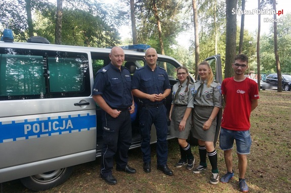 Zdjęcie wykonano w lesie. Przedstawia ono stojących na tle radiowozu policjantów i harcerzy.