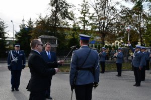 policjant składa meldunek, obok mężczyzna w czarnych okularach trzyma mikrofon, na przeciwko policjanta dwaj mężczyźni&quot;&gt;