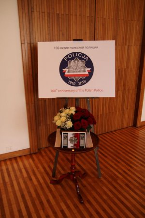Na stojaku plakat z logo 100-lecia powstania Policji Państwowej w języku polskim i rosyjskim, pod spodem bukiet biało - czerwonych kwiatów