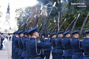 Policjanci plecami ustawieni do fotografa , mają karabiny na ramieniu z których uchodzi dym w tle wierza jasnogórska