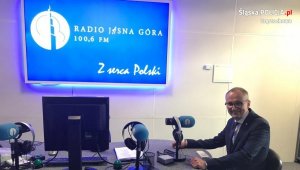 wiceminister spraw wewnętrznych i administracji siedzący za biurkiem na którym jest kilka mikrofonów oraz monitor w tle telebim reklamowy radia jasna góra