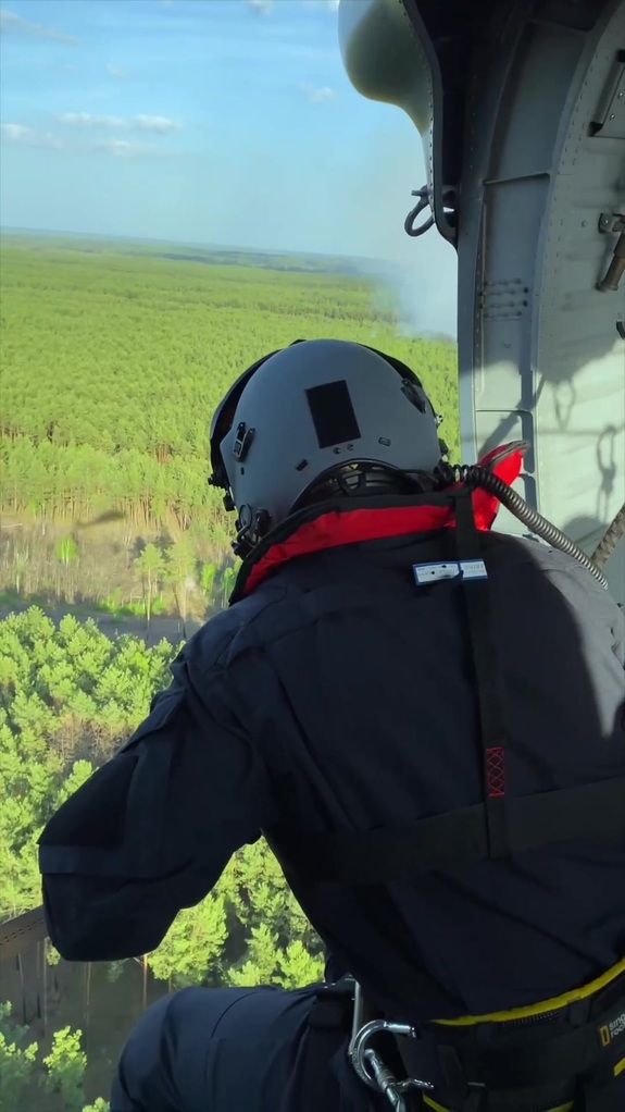 Mężczyzna w kasku siedzi w otwartych drzwiach helikoptera i obserwuje las.