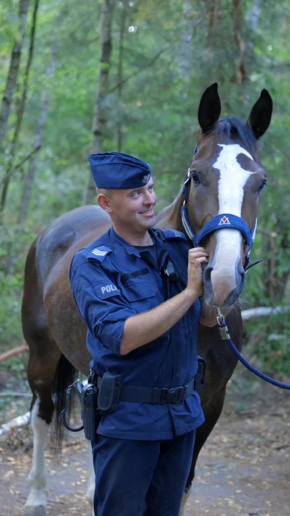 Uśmiechnięty policjant w furażerce trzyma za uzdę konia.