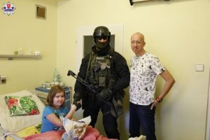 Policjanci i kontrterroryści z prezentami u małych pacjentów - policjant stoi przy łóżku chorej dziewczynki, obok policjanta stoi chory chłopiec