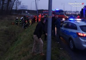 Na fotografii widać pobocze drogi, rów, w którym leży na boku samochód osobowy. Obok samochodu znajduje się kilku strażaków. Na drodze widać policyjny radiowóz oraz wozy strażackie