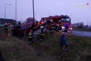 Na fotografii widać pobocze drogi, rów, w którym leży na boku samochód osobowy. Obok samochodu znajduje się kilku strażaków. Na drodze widać  wozy strażackie