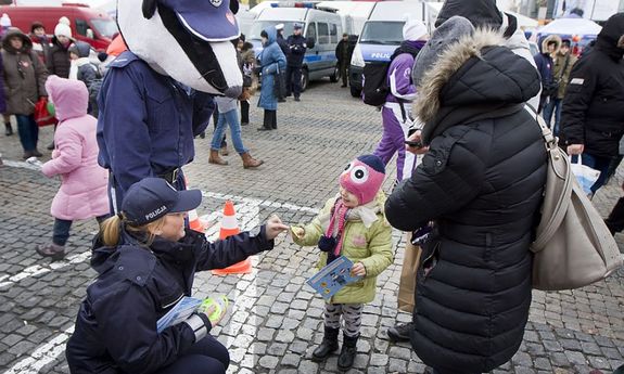 policjantka w mundurze wręcza element odblaskowy dziecku będącemu z mamą. Obok stoi policyjna maskotka - bobrek, z tyłu widoczni są policjanci, spacerujący ludzie i dwa policyjne radiowozy