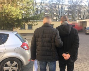 Na zdjęciu parking policyjny,  policjant prowadzi do radiowozu zatrzymanego mężczyznę