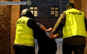 policjanci w kamizelkach odblaskowych z napisem policja prowadzą zatrzymanego w kajdankach