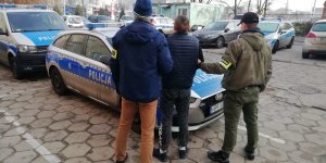 dwaj policjanci z zatrzymanym mężczyzną stoją przy radiowozie