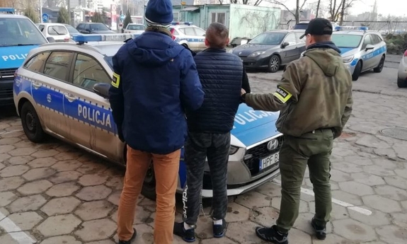 dwaj policjanci z zatrzymanym mężczyzną stoją przy radiowozie