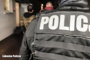 napis POLICJA na kamizelce policjanta, w tle widać zatrzymanych mężczyzn