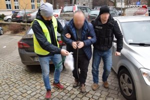 dwóch policjantów prowadzi zatrzymanego mężczyznę, który na dłoniach i stopach ma założone kajdanki zespolone