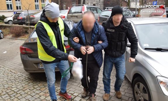 dwóch policjantów prowadzi zatrzymanego mężczyznę, który na dłoniach i stopach ma założone kajdanki zespolone&quot;&gt;