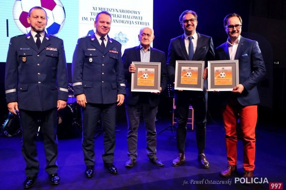 Zastępca Komendanta Głównego Policji, Komendant Stołeczny Policji, Andrzej Strejlaui oraz inni mężczyźni stoją na scenie trzymając w ręku pamiątki z turnieju podczas gali otwarcia XI Międzynarodowego Turnieju Służb Mundurowych w Piłce Nożnej Halowej im. podkom. Andrzeja Struja