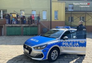 policjant stoi przy radiowozie, z tyłu, przy budynku grupa dzieci