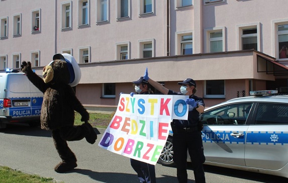 policyjny miś przy radiowozie i policjantki z plakatem „Wszystko będzie dobrze”