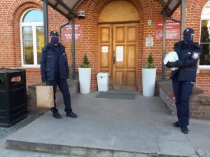 dwie policjantki w maskach na twarzach i z workami z maseczkami stoją przed budynkiem urzędu miasta