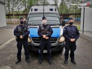 Policjanci z bielskiego pododdziału prewencji stoją przed radiowozem. Mundurowi mają zasłonięte twarze maseczkami