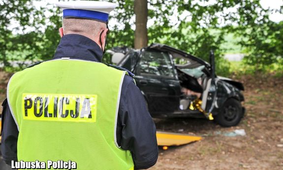 policjant w mundurze i kamizelce z napisem Policja na miejscu wypadku, z tyłu wrak rozbitego auta