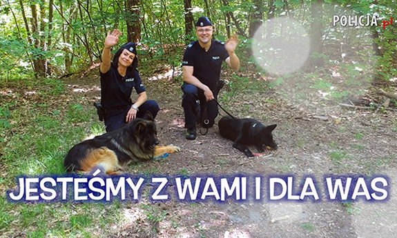 Para policjantów ze swoimi psami służbowymi macha do kamery. Napis: JEsteśmy z Wami i dla Was.