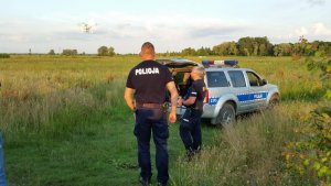 dwaj policjanci  w trakcie poszukiwań, w tle radiowóz policyjny i niskie zarośla, jeden z policjantów obsługuje drona latającego nad zaroślami