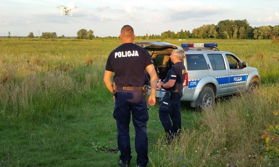 dwaj policjanci  w trakcie poszukiwań, w tle radiowóz policyjny i niskie zarośla, jeden z policjantów obsługuje drona latającego nad zaroślami