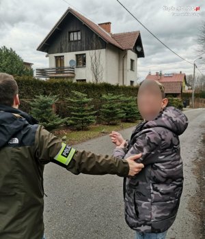 Na fotografii widać policjanta, który trzyma za rękę zatrzymanego mężczyznę. W oddali widać dom jednorodzinny.&quot;&gt;