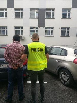 Na zdjęciu widać osobę zatrzymaną w kajdankach oraz policjanta na tle dwóch radiowozów nieoznakowanych.