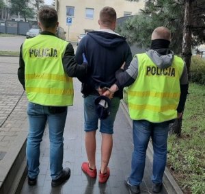 Zatrzymany stoi w krótkich spodenkach przy policyjnej izbie zatrzymań na dziedzińcu Komendy Miejskiej Policji w Łodzi, ma założone kajdanki z tyłu, po jego obu stronach stoi dwóch policjantów ubranych w żółte kamizelki.&quot;&gt;