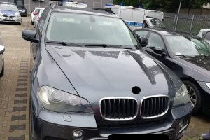 odzyskany przez policjantów samochód marki BMW X5, widok przodu pojazdu, który jest w kolorze ciemnym, w tle policyjne radiowozy i budynki