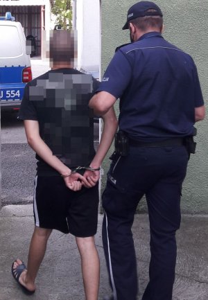 Policjant w mundurze trzyma za ramię mężczyznę, który ma założone kajdanki na ręce trzymane z tyłu