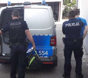 Dwóch policjantów w mundurach stoi przy otwartym radiowozie z tyłu.&quot;&gt;