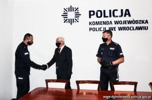 Na zdjęciu Wojewoda Dolnośląski Jarosław Obremski składa gratulacje podkomisarzowi Dariuszowi Wareckiemu w obecności Komendanta Wojewódzkiego Policji we Wrocławiu inspektor Dariusza Wesołowskiego w sali konferencyjnej Komendy Wojewódzkiej Policji we Wrocławiu na ścianie w tle nowe logo Policji - gwiazda policyjna