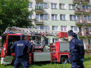 dwaj policjanci stoją przed blokiem mieszkalnym obok nich znajduje się wóz strażacki