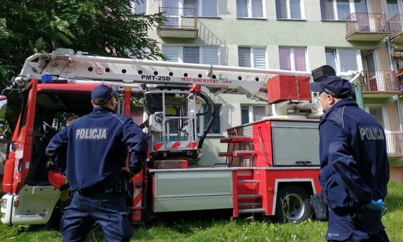 dwaj policjanci stoją przed blokiem mieszkalnym obok nich znajduje się wóz strażacki