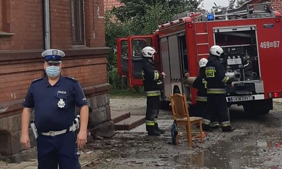 Policjant stoi koło budynku, z tyłu wóz straży pożarnej i strażacy