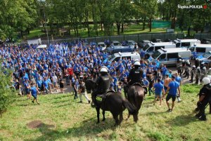 Na zdjęciu widoczna grupa osób, oznakowane radiowozy policjanci w kaskach, kamizelkach trzymający tarcze oraz policja konna.&quot;&gt;