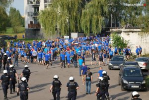 Na zdjęciu widoczna grupa osób, która idzie w kierunku bloku, za nimi idą policjanci w kaskach i kamizelkach.&quot;&gt;